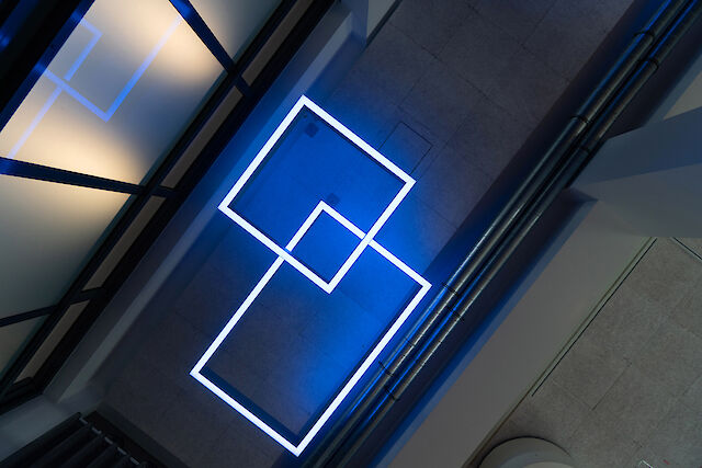 View from above of two intertwined, luminous squares of light on the ceiling of a modern interior. The geometric luminaires shine in clear blue and stand out against the grey floor and the visible installation pipes