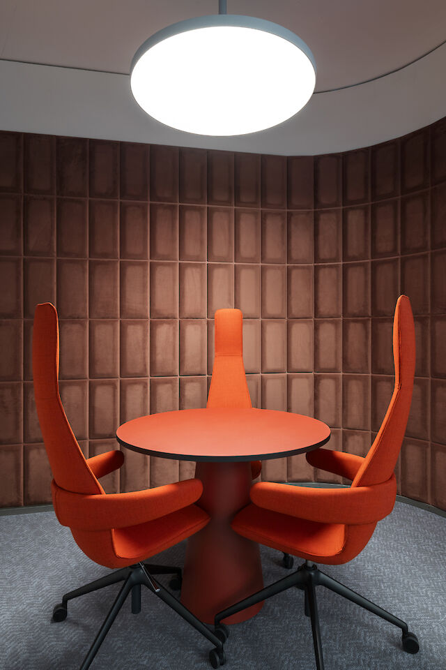 Runder Besprechungstisch mit drei hohen roten Stühlen unter runder Pendelleuchte vor brauner Wand