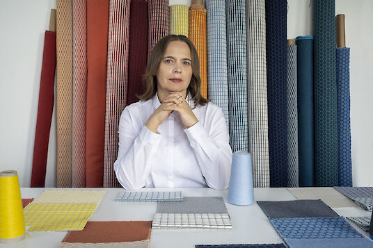Textile designer Aleksandra Gaca sits at a table with fabric samples, surrounded by colourful rolls of acoustic textiles.